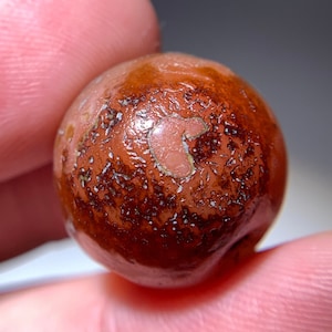 Puede incluir: Primer plano de una piedra redonda de color marrón rojizo con una superficie texturizada. La piedra tiene un área de color más claro con un patrón único. La piedra se sostiene entre dos dedos.