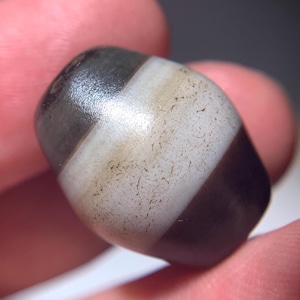 Puede incluir: Una cuenta de ágata negra y blanca con una superficie lisa y pulida. La cuenta tiene tres rayas blancas y es de forma ovalada.