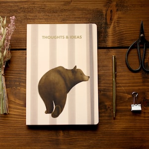 May include: A white and grey striped notebook with a brown bear illustration and the text "THOUGHTS & IDEAS" on the cover. The notebook is on a wooden surface with a bunch of dried flowers, a pair of scissors, a pen, and a binder clip.