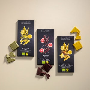 Könnte beinhalten: Drei Schokoladentafeln mit verschiedenen Geschmacksrichtungen: Matcha-Zitrone, Chia-Grapefruit und Kurkuma-Ingwer-Zitrone. Jede Tafel hat eine andere Farbe und zeigt eine Fruchtillustration. Die Tafeln sind biologisch und vegan.