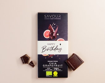 May include: A dark chocolate bar with a pink grapefruit and chia seeds on the front. The bar is wrapped in black and white packaging with the words "Happy Birthday" and "Savour Finest Experience". The bar is organic, vegan, and 70% cacao.