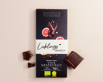 May include: A dark chocolate bar with a black and white label that says "SAVOUR FINEST EXPERIENCE" and "Lieblings MENSCH". The label also says "ORGANIC | VEGAN CHIA GRAPEFRUIT DARK CACAO BAR 70% CACAO".