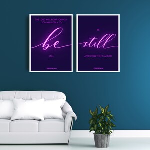 May include: Two framed prints with white frames and a dark purple background. The text on the left print reads "THE LORD WILL FIGHT FOR YOU YOU NEED ONLY TO be still STILL - EXODUS 14:14". The text on the right print reads "BE still AND KNOW THAT I AM GOD - PSALMS 46:10". The text is in a pink neon font.
