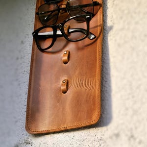 Puede incluir: Un organizador de pared de cuero marr&oacute;n con tres ganchos para colgar gafas. Dos pares de gafas est&aacute;n colgados de los ganchos.