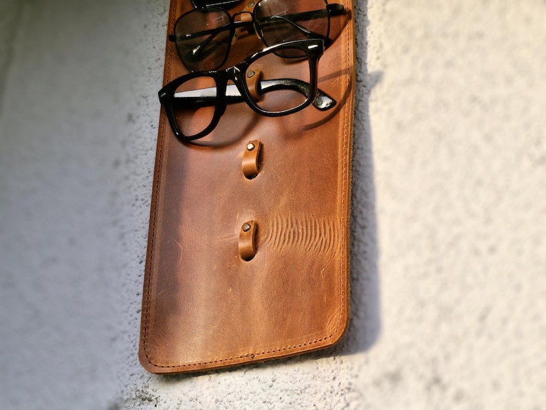 Puede incluir: Un organizador de pared de cuero marr&oacute;n con tres ganchos para colgar gafas. Dos pares de gafas est&aacute;n colgados de los ganchos.