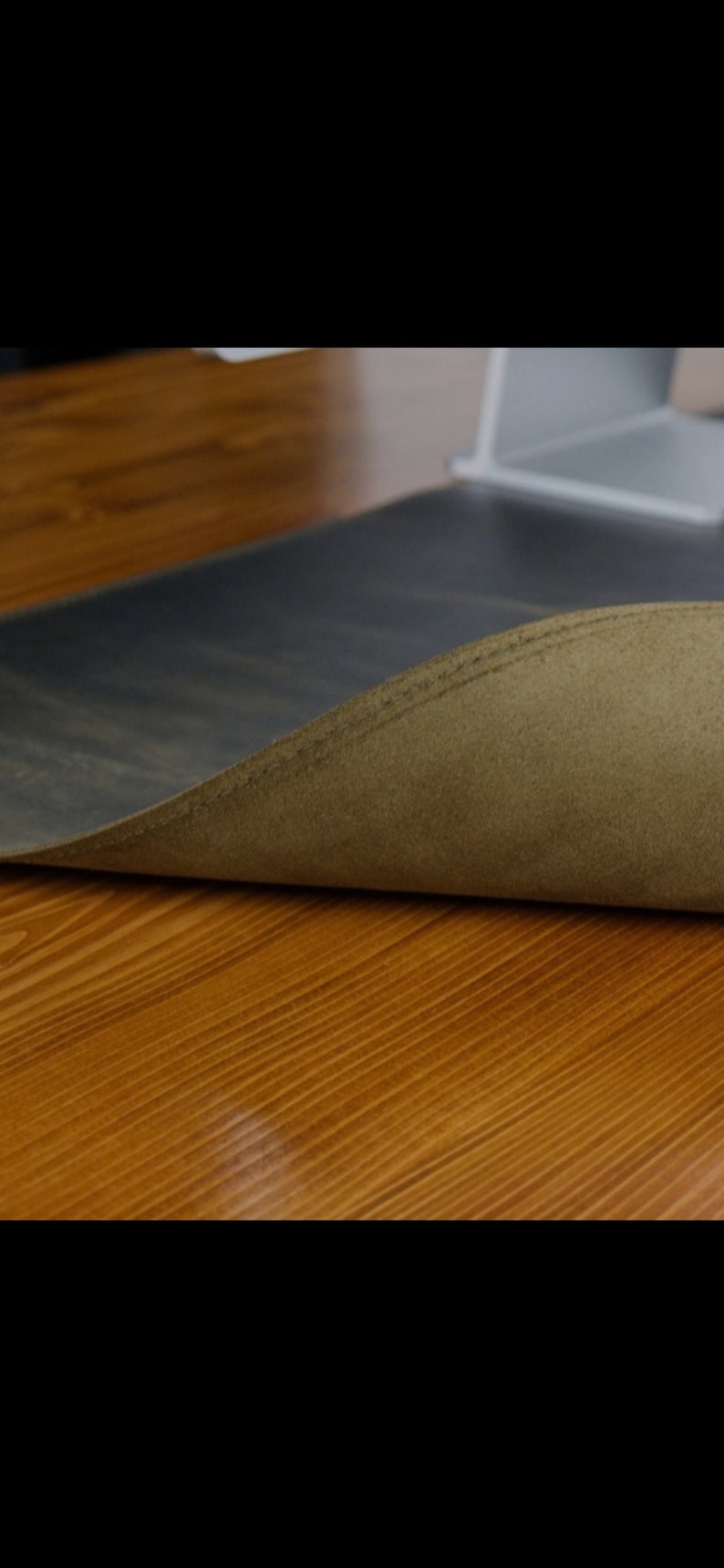 May include: A dark charcoal gray desk mat with a smooth top surface and a textured, olive-toned suede underside. The mat is slightly curled at the edge, revealing the contrast between the dark upper material and the lighter, fuzzy bottom layer.