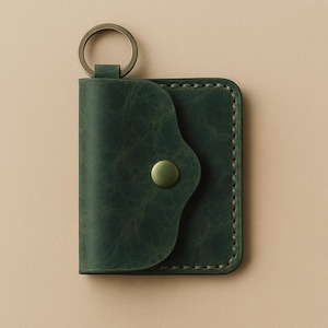 May include: A dark green leather key holder with a metal key ring. The key holder has a curved flap secured by a brass-colored snap. The edges are stitched with a matching green thread, and the leather has a textured appearance.