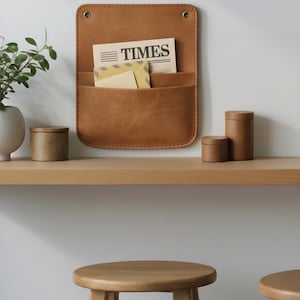 May include: A tan leather wall-mounted organizer with two pockets containing a newspaper with the text TIMES and envelopes. The rectangular organizer features rounded corners, visible stitching along the edges, and two metal grommets at the top.