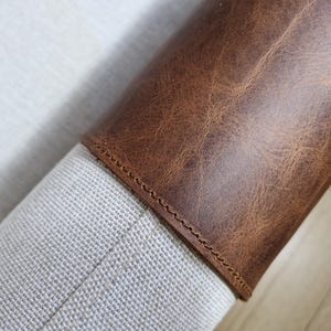 May include: A cylindrical object featuring a textured, medium-brown leather wrap secured with visible stitching. The leather section is attached to a light-colored, woven fabric base that has a visible, fine-grained, off-white texture.
