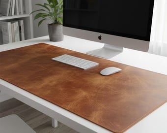 May include: A brown leather desk mat with a distressed finish, a white keyboard, and a white mouse sit on a white desk. A computer monitor and a potted plant are also on the desk. The desk is in front of a white bookshelf.