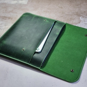 Peut inclure: Étui pour ordinateur portable en cuir vert avec intérieur vert vif. L'étui rectangulaire a des coins arrondis et des coutures blanches. Un ordinateur portable argenté est partiellement visible. Deux fermetures à pression sécurisent le rabat.