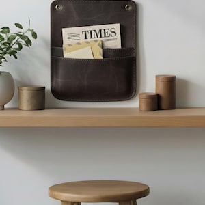 May include: A dark brown leather wall-mounted organizer with two pockets, featuring two brass-colored metal grommets at the top. The organizer holds a newspaper with the text TIMES and a light-colored envelope tucked into the front pocket.