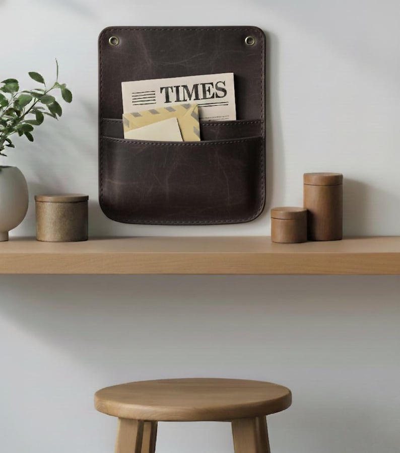 May include: A dark brown leather wall-mounted organizer with two pockets, featuring two brass-colored metal grommets at the top. The organizer holds a newspaper with the text TIMES and a light-colored envelope tucked into the front pocket.
