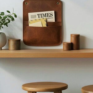May include: A brown leather wall-mounted organizer with two pockets, containing a newspaper with the word TIMES printed on it and a yellow envelope. The organizer features two brass-colored metal grommets at the top corners for mounting.