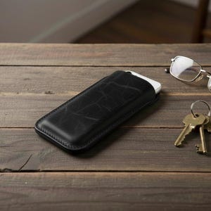 May include: A black leather phone case with a white phone inside, resting on a wooden surface. Also present are a pair of eyeglasses and a set of keys.