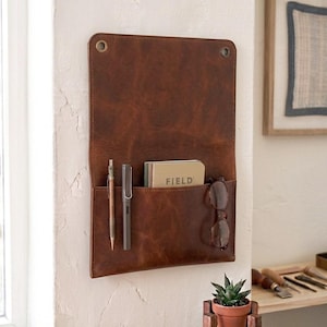 May include: A rectangular brown leather wall organizer featuring two metal grommets at the top corners for mounting. The front pocket holds a metallic mechanical pencil, a grey fountain pen, a notebook labeled FIELD, and a pair of brown eyeglasses.