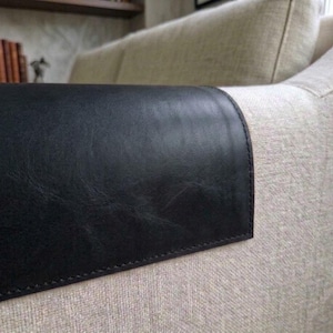 May include: A black leather armrest cover draped over the beige fabric arm of a sofa. The cover features visible stitching along the edges and a smooth, slightly textured surface that contrasts with the woven light-colored upholstery.