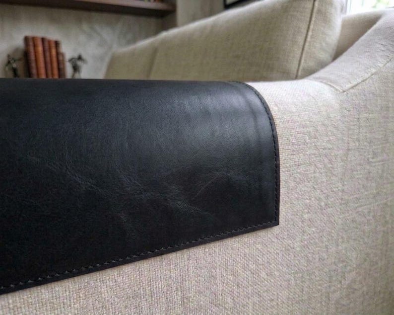 May include: A black leather armrest cover draped over the beige fabric arm of a sofa. The cover features visible stitching along the edges and a smooth, slightly textured surface that contrasts with the woven light-colored upholstery.