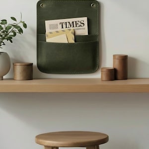 May include: A dark olive green wall-mounted organizer with two pockets, featuring brass-toned eyelets at the top. The organizer holds a newspaper with the word TIMES printed on it and a light-colored envelope tucked into the front pocket.