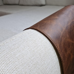 May include: A dark brown leather armrest cover draped over the light beige fabric arm of a sofa. The leather features a smooth, slightly distressed texture with visible stitching along the curved edges, fitting snugly over the rounded furniture arm.