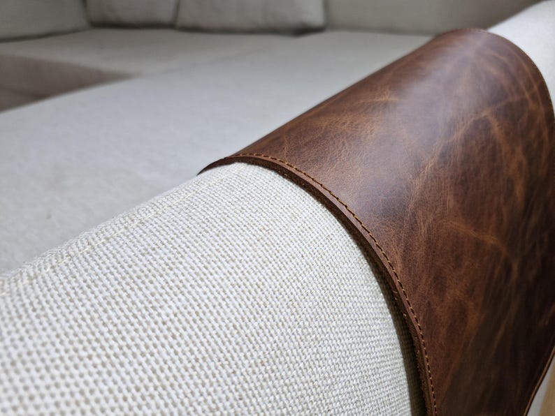 May include: A dark brown leather armrest cover draped over the light beige fabric arm of a sofa. The leather features a smooth, slightly distressed texture with visible stitching along the curved edges, fitting snugly over the rounded furniture arm.