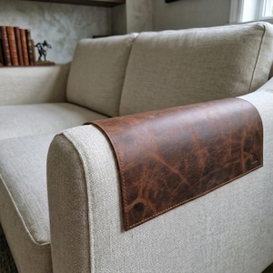 May include: A rectangular brown leather armrest cover with a distressed texture and visible stitching along the edges, draped over the arm of a light beige fabric sofa. The leather piece features a small embossed logo on the lower right corner.
