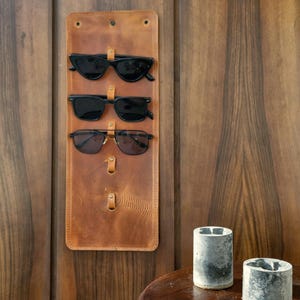 Puede incluir: Un soporte de gafas de sol de cuero marr&oacute;n con tres pares de gafas de sol colgadas. El soporte est&aacute; montado en una pared de madera.