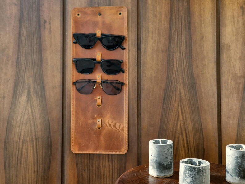 Puede incluir: Un soporte de gafas de sol de cuero marr&oacute;n con tres pares de gafas de sol colgadas. El soporte est&aacute; montado en una pared de madera.
