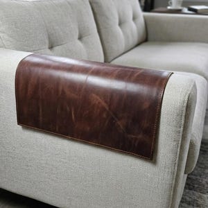 Puede incluir: Una funda rectangular de cuero marrón oscuro para reposabrazos colocada sobre el brazo de un sofá tapizado en color beige claro. El cuero presenta un acabado liso y ligeramente desgastado con grano natural visible y bordes cosidos.
