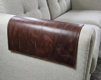 May include: A rectangular dark brown leather armrest cover draped over the arm of a light beige upholstered sofa. The leather features a smooth, slightly distressed finish with visible natural grain and stitched edges along the perimeter.