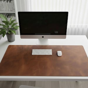 May include: A brown leather desk mat with a computer, keyboard, and mouse. The desk mat is rectangular and covers most of the white desk surface. A potted plant and a bookshelf are visible in the background.