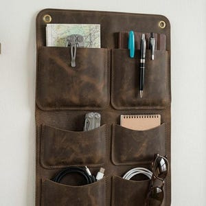 May include: A dark brown leather wall organiser featuring six individual pockets arranged in two columns. The pockets contain various items including a map, a multi-tool, pens, a small notebook, charging cables, and a pair of sunglasses.