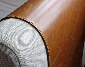 May include: A close-up view of a furniture armrest featuring a smooth, tan-colored leather panel attached to a textured, off-white woven fabric section. The leather is secured to the fabric with a single row of precise, tonal stitching along the edge.