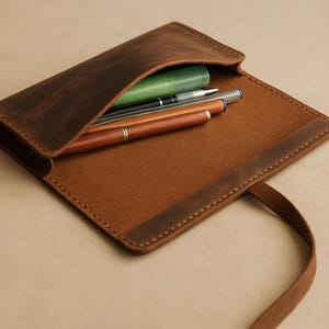 Peut inclure: Étui à stylos en cuir marron ouvert, révélant plusieurs stylos et un surligneur vert. L'étui a un rabat et une sangle. Le cuir a une finition vieillie et les coutures sont visibles.