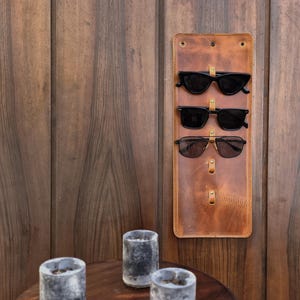 Puede incluir: Un soporte para gafas de sol de cuero marr&oacute;n con tres pares de gafas de sol negras colgadas. El soporte est&aacute; montado en una pared de madera.