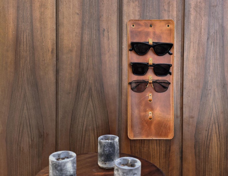 Puede incluir: Un soporte para gafas de sol de cuero marr&oacute;n con tres pares de gafas de sol negras colgadas. El soporte est&aacute; montado en una pared de madera.