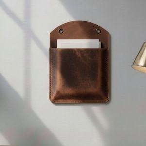 May include: A dark brown leather wall-mounted pocket organiser with a rounded top flap and two metal grommets. The rectangular pouch features visible stitching along the edges and contains several white rectangular papers tucked inside the main compartment.