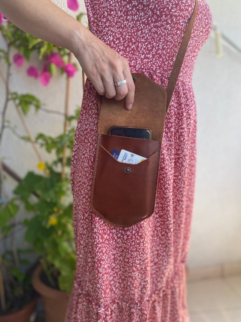 May include: A brown leather crossbody phone bag with a flap closure. The bag is worn over a red floral dress.