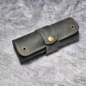 Puede incluir: Un estuche enrollable de cuero gris oscuro con cierre de bot&oacute;n de lat&oacute;n. El estuche est&aacute; hecho de cuero texturizado y tiene una costura cosida alrededor de los bordes.