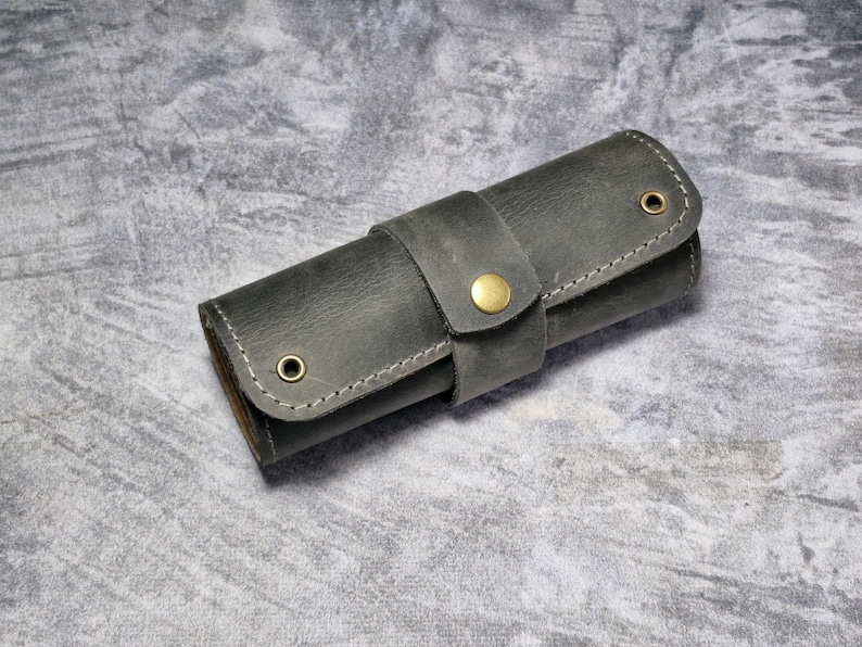 Puede incluir: Un estuche enrollable de cuero gris oscuro con cierre de bot&oacute;n de lat&oacute;n. El estuche est&aacute; hecho de cuero texturizado y tiene una costura cosida alrededor de los bordes.