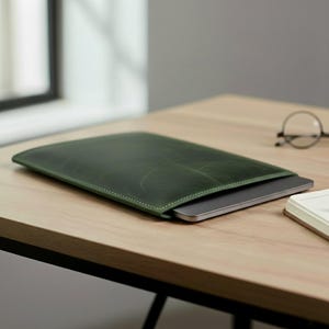 以下が含まれることがあります： 白いステッチが施された濃い緑色の革製ラップトップスリーブ。 スリーブは開いており、内側にシルバーのラップトップが現れています。 スリーブは明るい木材の表面にあります。