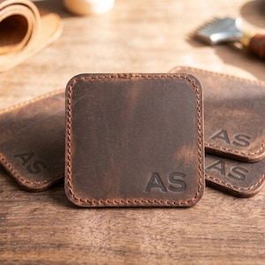 May include: A set of square brown leather coasters with rounded corners and visible stitching along the edges. Each coaster features the embossed letters AS in the bottom right corner. The leather has a matte, textured finish with natural variations.