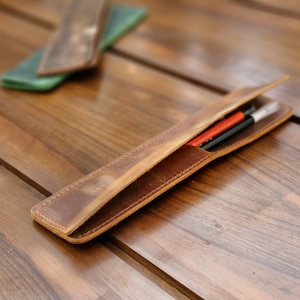 May include: A brown leather pencil case with a flap closure. The case is open and contains two pencils, one red and one black.