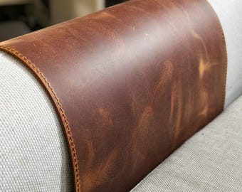 May include: A smooth, dark brown leather armrest cover draped over a light gray fabric sofa. The leather features visible natural grain variations and is secured with a consistent, decorative stitched border along the curved edges.