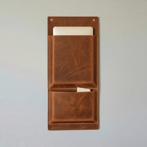 May include: A vertical brown leather wall organiser featuring two stacked pockets with visible stitching and brass-toned hardware at the top corners. A white rectangular electronic device is partially inserted into the top pocket and a small white object in the bottom.