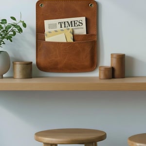 May include: A rectangular brown leather wall organizer with rounded corners, two pockets, and two brass-colored metal grommets at the top. The pockets contain a newspaper with the word TIMES printed on it and a light-colored envelope.