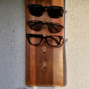 Puede incluir: Un soporte para gafas de cuero marr&oacute;n con tres pares de gafas colgadas. Las gafas son todas negras y tienen forma rectangular.