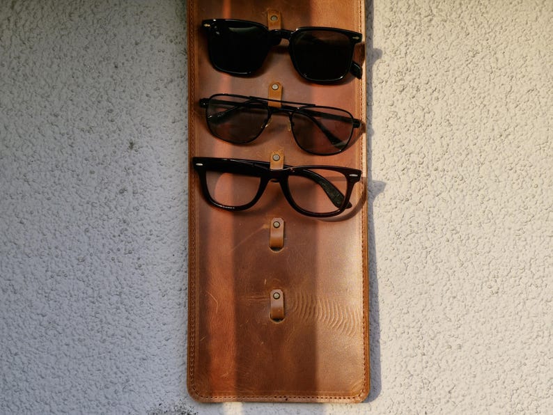 Puede incluir: Un soporte para gafas de cuero marr&oacute;n con tres pares de gafas colgadas. Las gafas son todas negras y tienen forma rectangular.