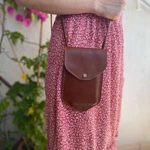 May include: A brown leather crossbody bag with a flap closure and a small pocket on the front. The bag is worn over a red floral dress.