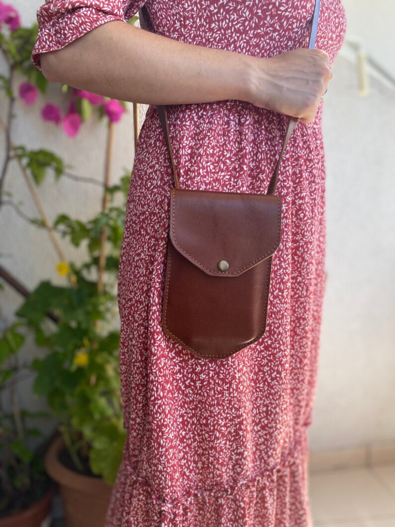 May include: A brown leather crossbody bag with a flap closure and a small pocket on the front. The bag is worn over a red floral dress.
